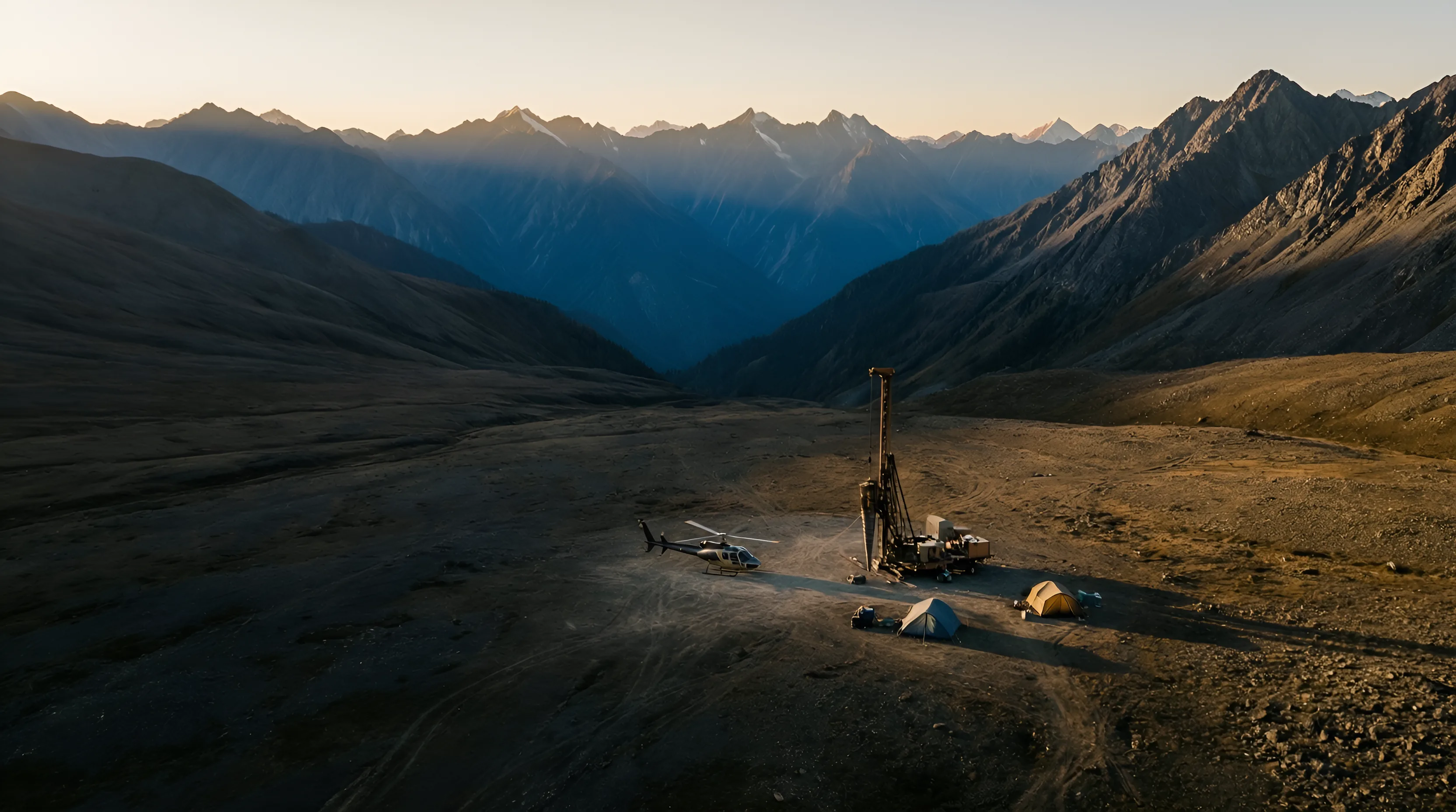 A lone drill rig, helicopter and field tents dwarfed by a vast remote mountain valley at dusk